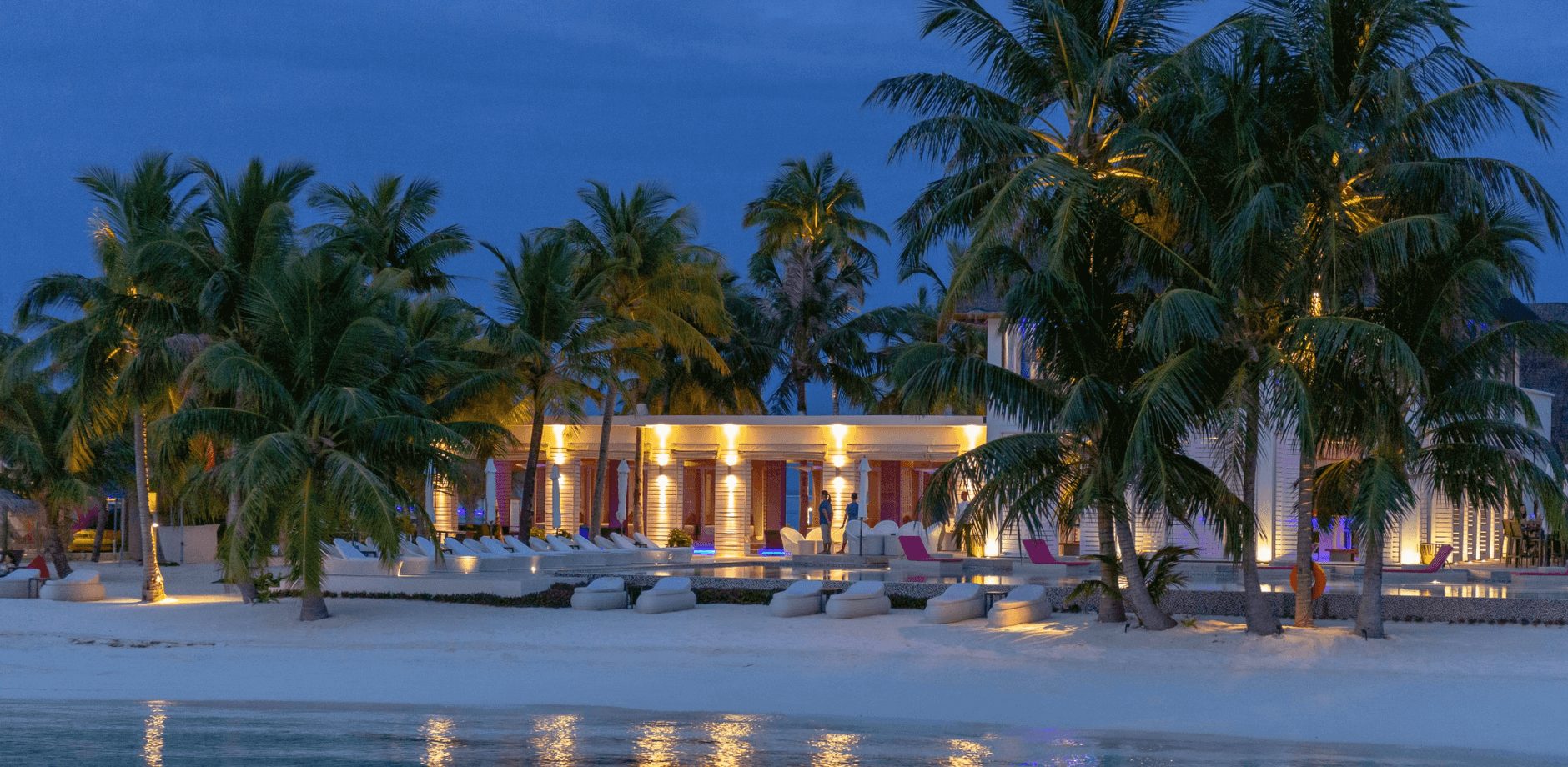 Beachfront resort at dusk with lit palm trees, loungers on white sand and a calm lagoon